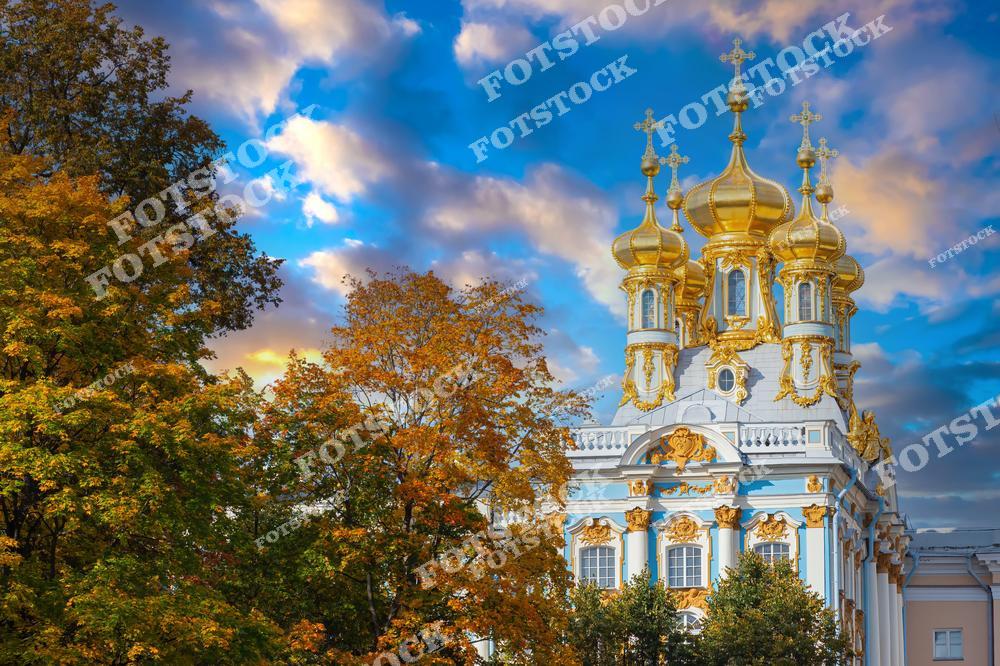На фотографии изображена церковь Воскресения Христова Екатерининского дворца в Пушкине (Царское Село). Золотые купола сверкают на фоне яркого неба, а осенние деревья добавляют красок и уюта в композицию. Это уникальный пример русской барочной архитектуры, расположенный в живописном парке. Здание является одним из символов величия императорской России.